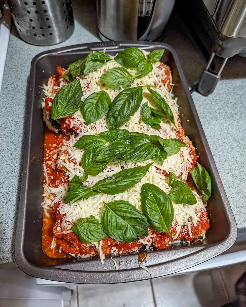 a not yet cooked eggplant parmesan in a 13x9 pan. there are fresh basil leaves atop a layer of cheese atop more layers of eggplant and cheese and marinara