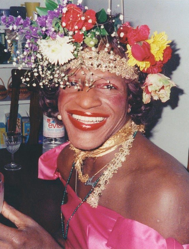 Photo of Marsha P Johnston, a smiling black woman in a pink dress with a flower crown on her head. 