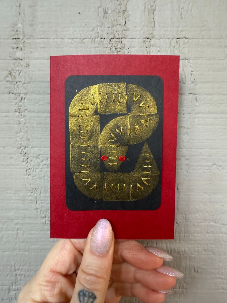 A hand holds a red card that has a black artist trading card on it. The artist trading card features. A custom rubber stamp of a snake curling in on itself. The snake is in gold metallic ink. The snake is stitched with gold thread and has red eyes sewn with embroidery floss.