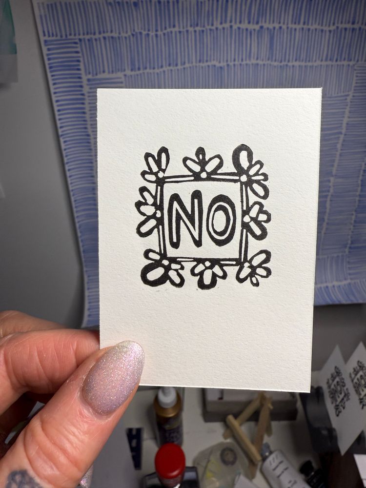 A hand holds a small piece of white paper with a linocut of the word “no”
