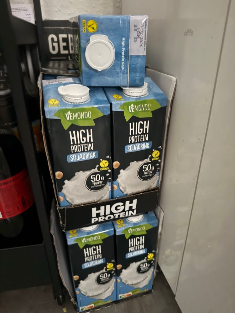 Mehrere Packungen Vemondo High Protein Sojadrink, gestapelt in Kisten. Die Packung ist hauptsächlich schwarz, mit blau gemischt und es steht ein schwarzer Button überall drauf mit 50g Protein pro Packung . Darüber in grün das Vemondo Logo von Lidl. 
