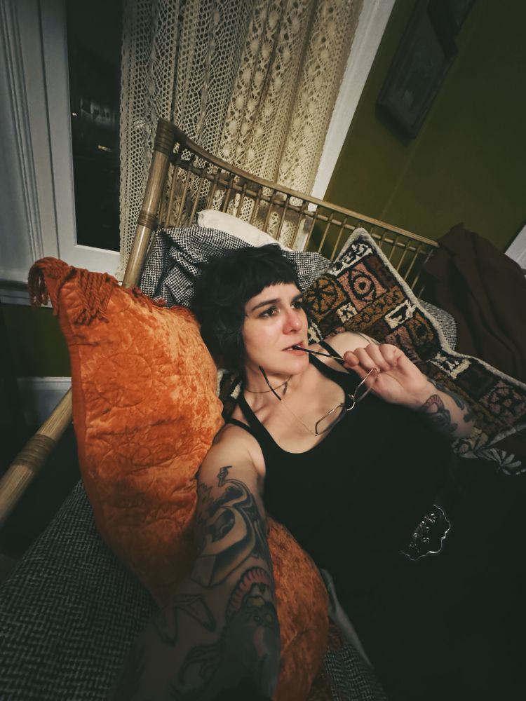 Photo of a selfie of a person in all black, leaning on cushions with their glasses in their mouth like a failed coquettish idiot  