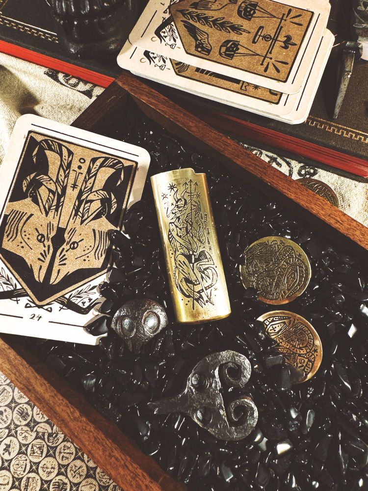 Photo of charms, including a divination card, etched coins, etched lighter cover, iron devil, iron skull in a tray of black gemstones 