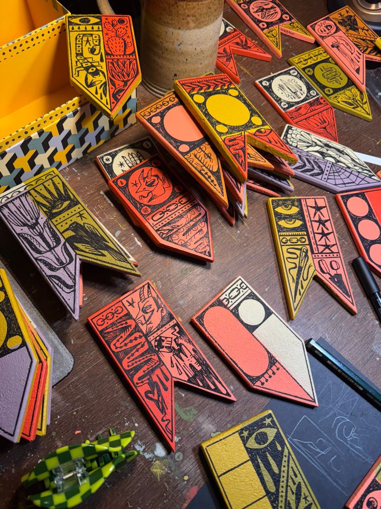 Photo of a slew of wooden pennants painted and drawn on in various states of finished. The gritty texture applied in the paint gives for good effects on some but caused sloppy drawings on others 