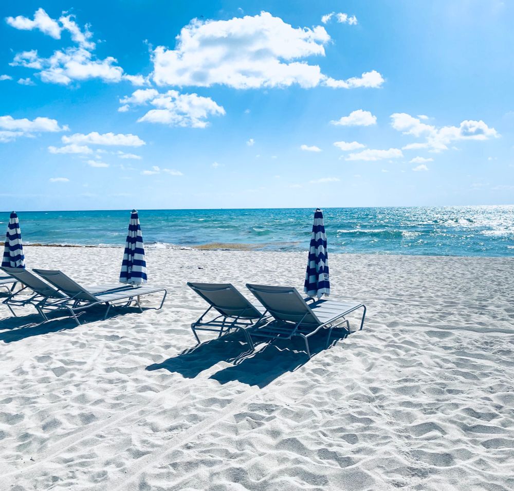 A sunny, blue, cloud sprinkled sky, above a green blue ocean at the beach. A few chaise lounges and umbrellas are positioned on the sand 