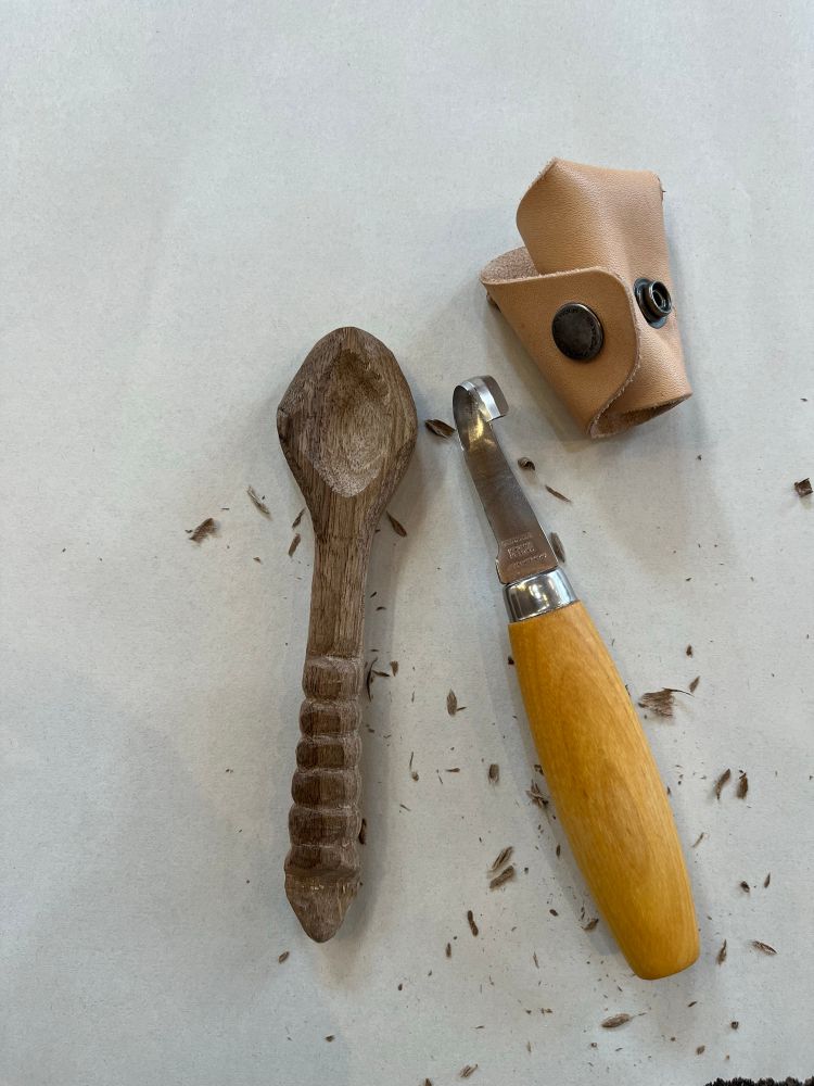 A walnut spoon with a curved, ribbed handle and a diamond shaped bowl sits next to a Morakniv spoon knife.