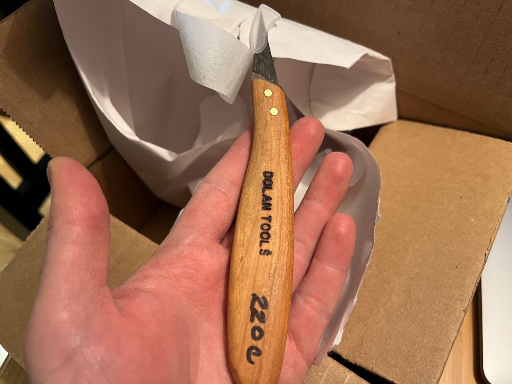 Will’s hand is holding a Dolan Tools 220c flexible blade knife with a cherry handle over a cardboard box with visible packing material