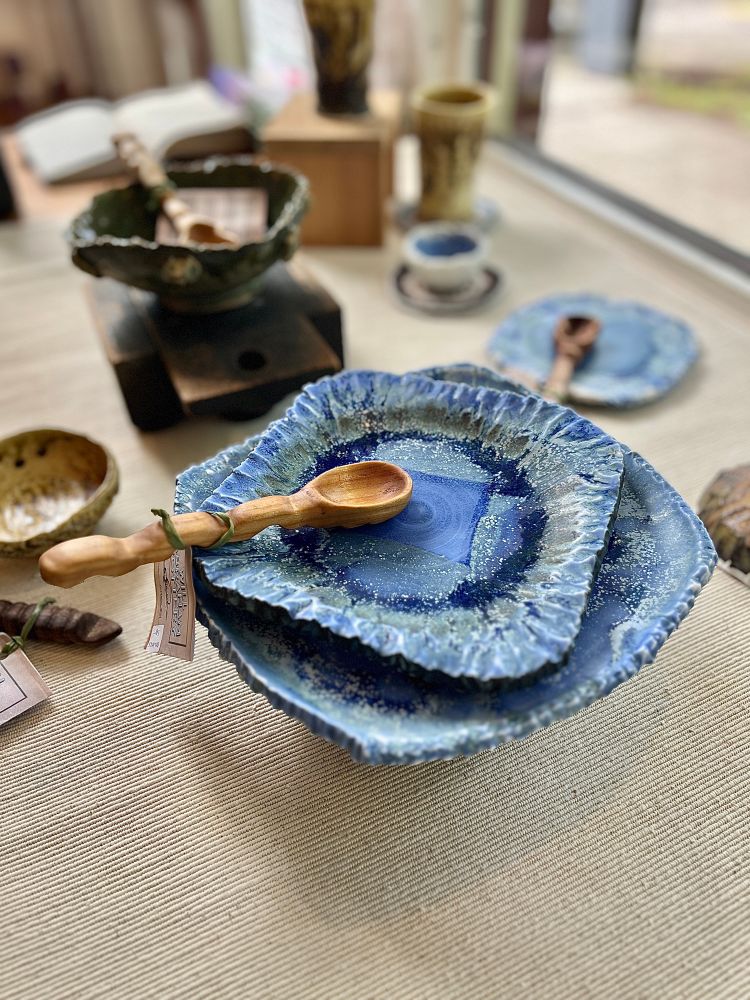 A stack of blue plates with rocky rims and crystals on the surface sit under a cherry spoon with a wiggly handle. Other tableware with similar motifs is visible in the background.