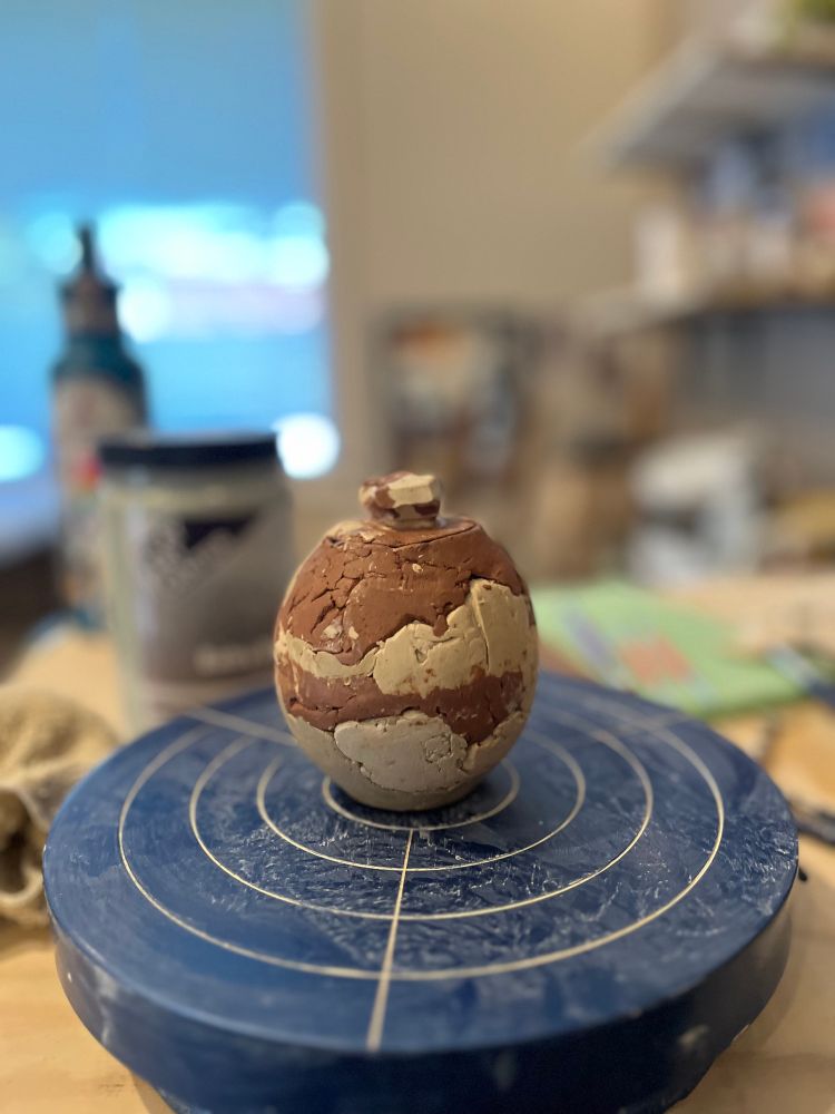 A 2.5 inch tall greenware jar with a lid with a small angular knob. Its surface has a rocky texture made out of black, white, grey, and yellow clays sitting in the center of a blue banding wheel. 
