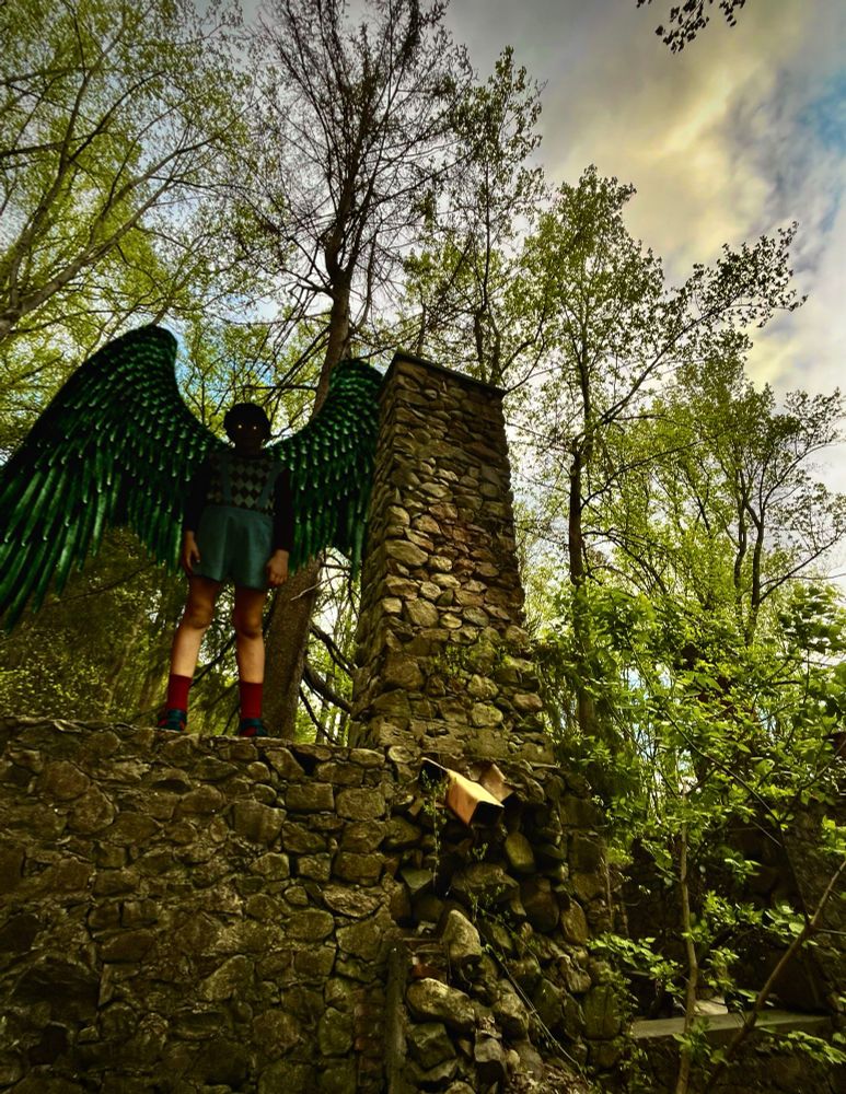 A winged child with no face and yellow eyes sits on a stone wall in the forest.