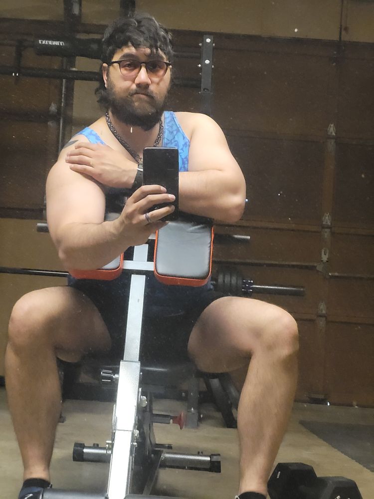 Masculine presenting person sitting on workout bench with a preacher pad attachment. Showing off biceps 