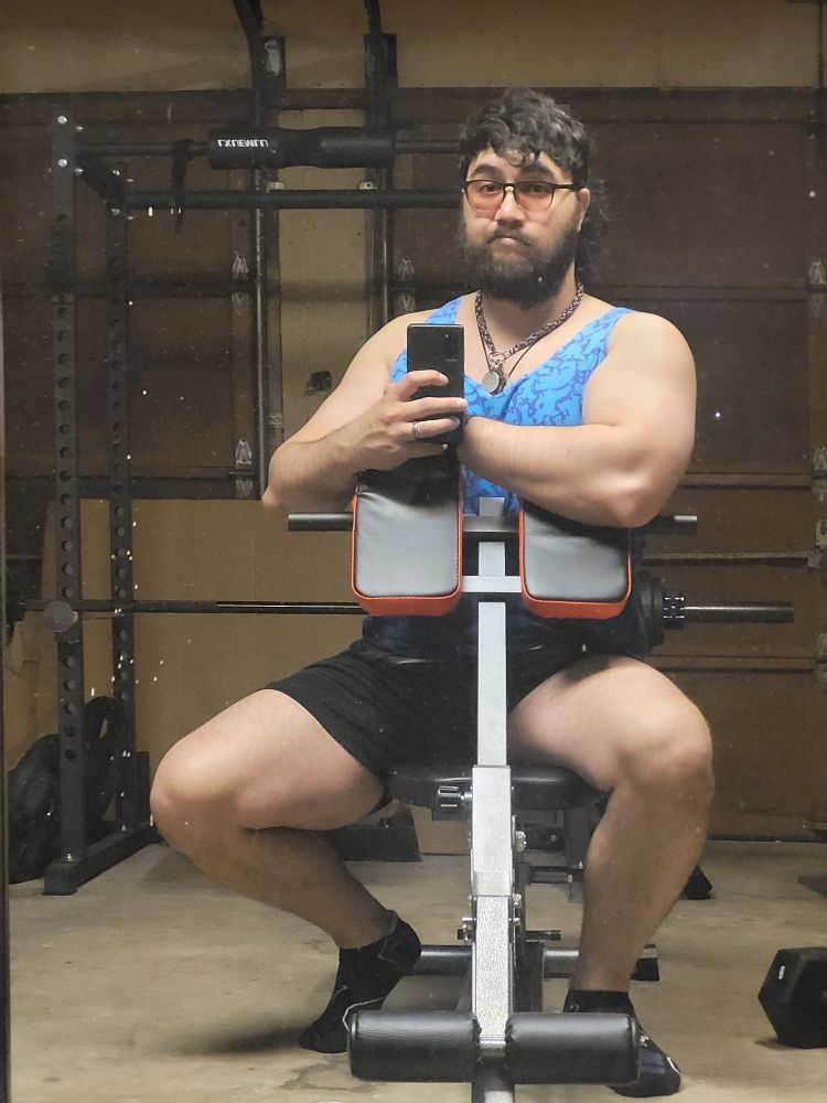 Masculine presenting person sitting on workout bench with a preacher pad attachment. Showing off biceps