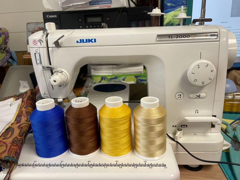 4 ounce cones of bright blue, medium brown, yellow, and off white thread sitting on the bed extension of my not-quite-industrial Juki TL-2000Qi sewing machine, which is sitting on one end of my desk. Off to the left are canvas and upholstery fabric waiting to be sewn with the thread.