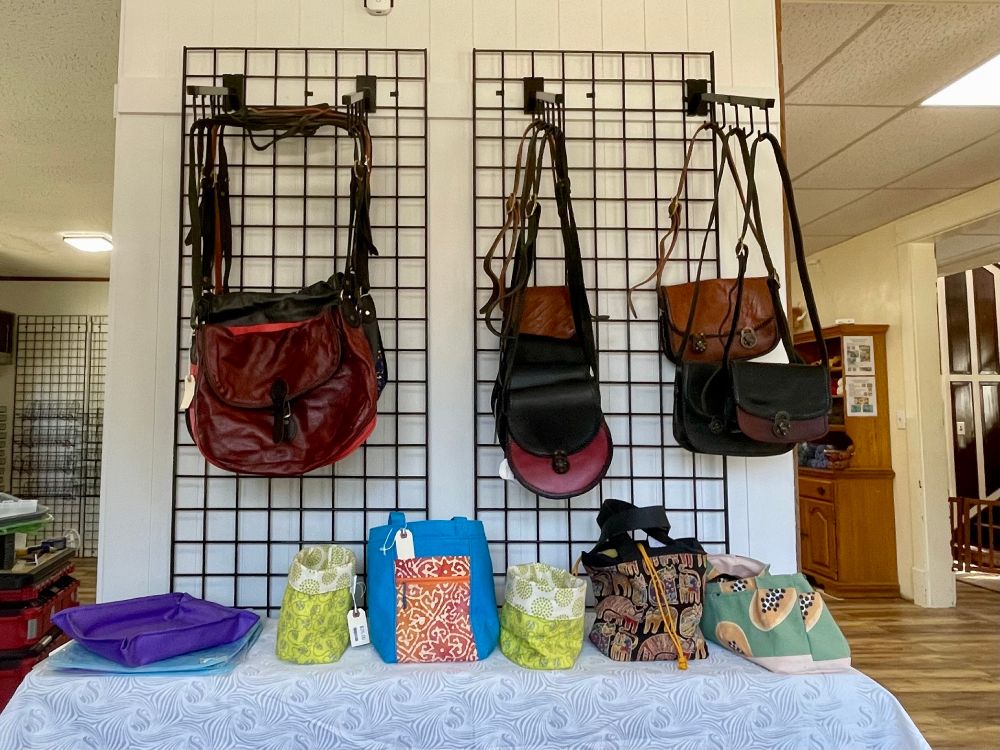 Leather bags and purses hanging from waterfall displays on black gridwall mounted to a white wall. Before the hanging bags are tote- and tray-type bags on a narrow table covered with a white-on-white cloth covered with swirly patterns. Other bits of the shop are visible on either side.