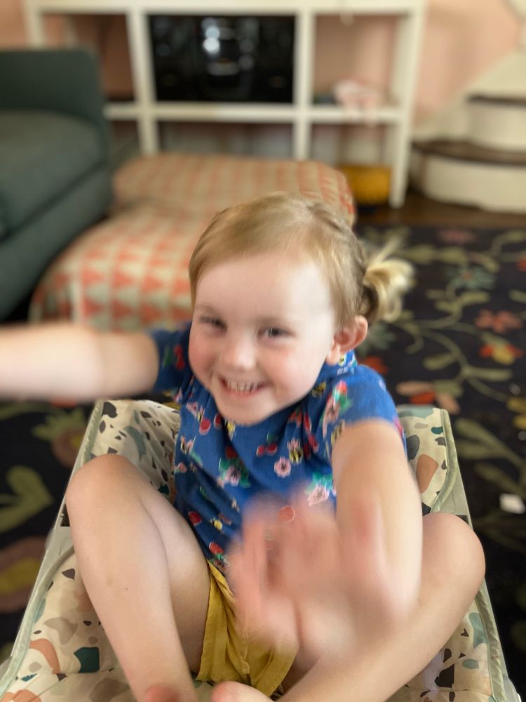 photo interior day. a blurred, smiling blonde child with pigtails reaches for the camera.