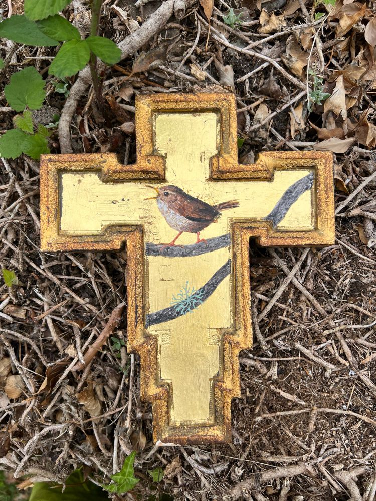 A gold cross with a wren perched on a bough is painted on a gold leaf background. It lays upon the ground which is scattered with twigs 