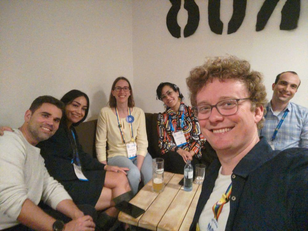 A cheerful group of individuals seated around a coffee table, all smiling at the camera, in a cosy setting.