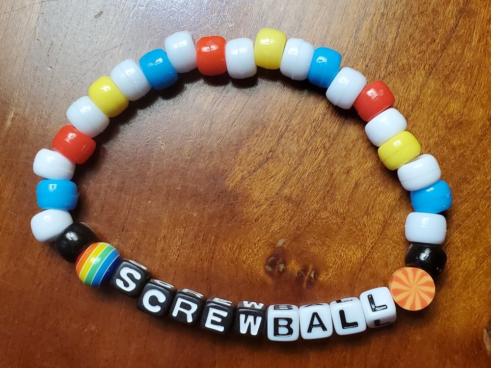 A kandi bracelet made primarily of pony beads. The center says "Screwball" with the "screw" in black blocks with white letters and "ball" in white blocks with black letters. To the left of "screw" is a spherical bead with rainbow stripe, and to the left of that ball is a black pony bead. To the right of "ball" is flat orange circular bead with a spiral, and to the right of that is another black pony bead. The rest of the bracelet alternates between white and then either blue, red, or yellow.