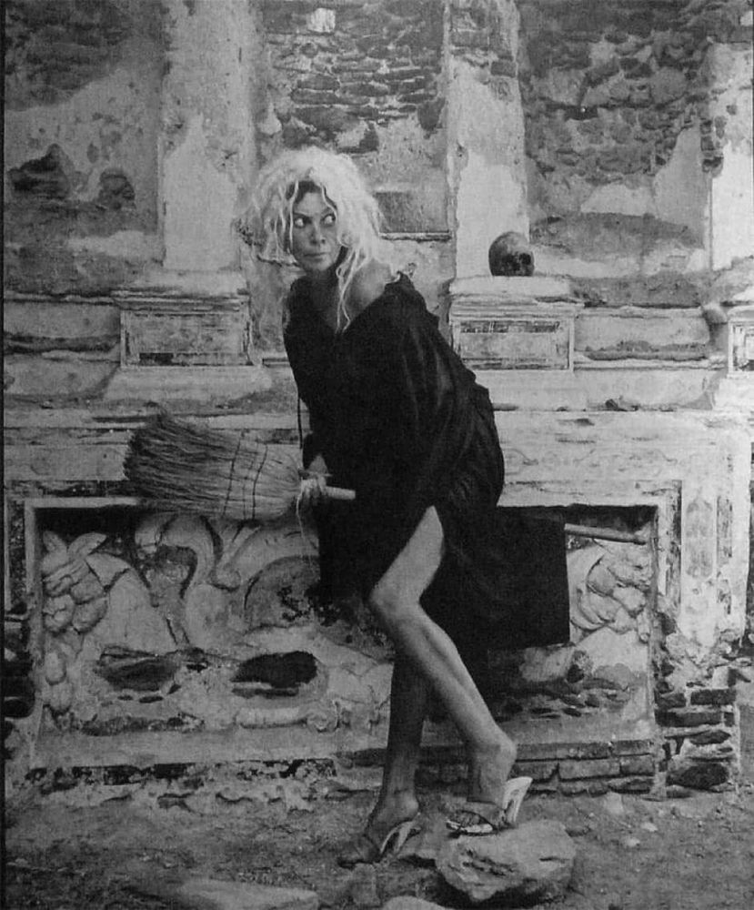 A witchy photograph of artist Leonor Fini, straddling a broomstick, photographed in a derelict and crumbling Corsican monastery. She is wearing a white/blonde wig and a shapeless black dress that exposes her thigh, paired with heels. She is giving epic side-eye- peering naughtily and mischievously behind her.