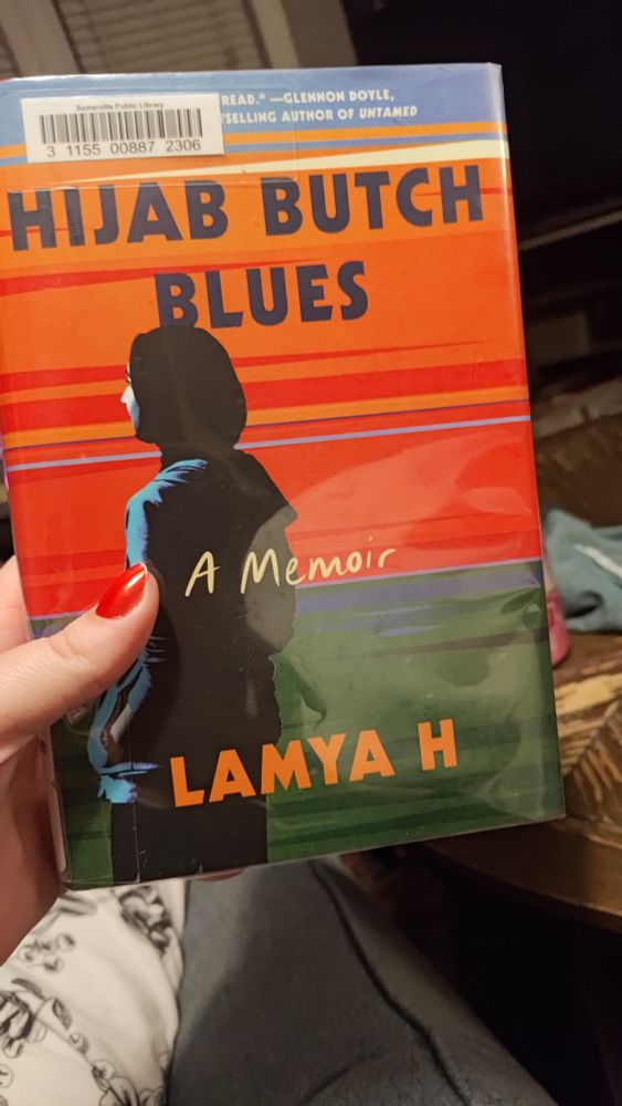 A hand holding the book HIJAB BUTCH BLUES by Lamya H. The cover is a woman turned three quarters away from us wearing a hijab and hoodie sweatshirt with her hands in her pockets. She is looking at a graphic that is orange, red, blue, and green, reminiscent of a sunset over the water.