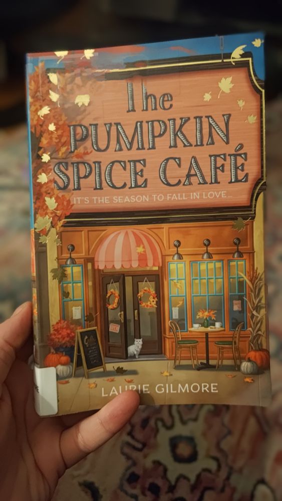 A hand holding the book The Pumpkin Space Cafe by Laurie Gilmore. The cover shows a drawing of a storefront with a bistro table for two in front; on the table are two mugs and a plate with a pastry. The double doors are covered with a red and pink striped awning with scallops; autumnal wreaths hang on the glass doors. One of the doors is ajar and a white cat is coming out. Decorative pumpkins, florals, and corn stalk are arranged around the cafe front.
