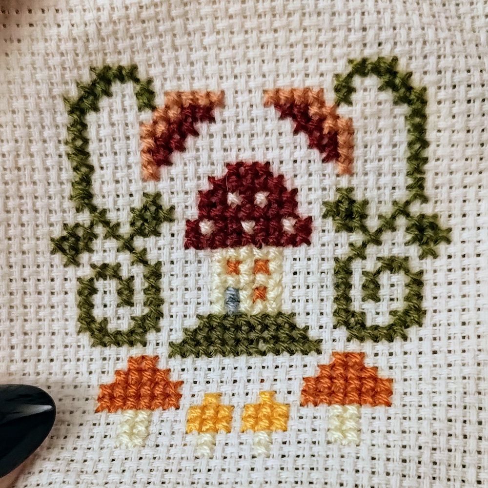 A 3x3 inch cross stitch of a mushroom cottage. There are 4 smaller mushrooms below it, and on either side is a flower with a swirly stem.