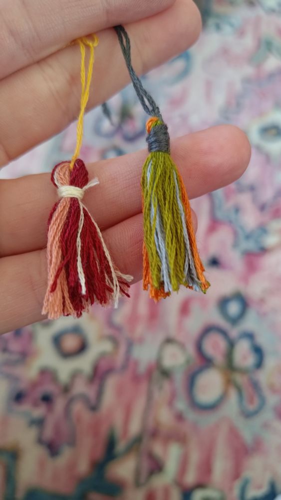 A hand holding two small tassels. The tassels are about two finger-widths long. One is maroon and blush; the other is moss, orange, and sky blue.