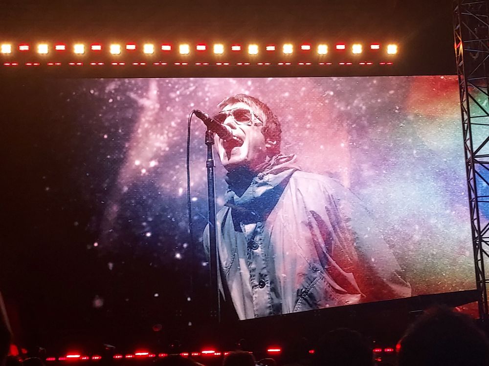 Liam Gallagher wearing sunglasses and a raincoat, singing, as seen on video screen during 12 September concert in Mexico City