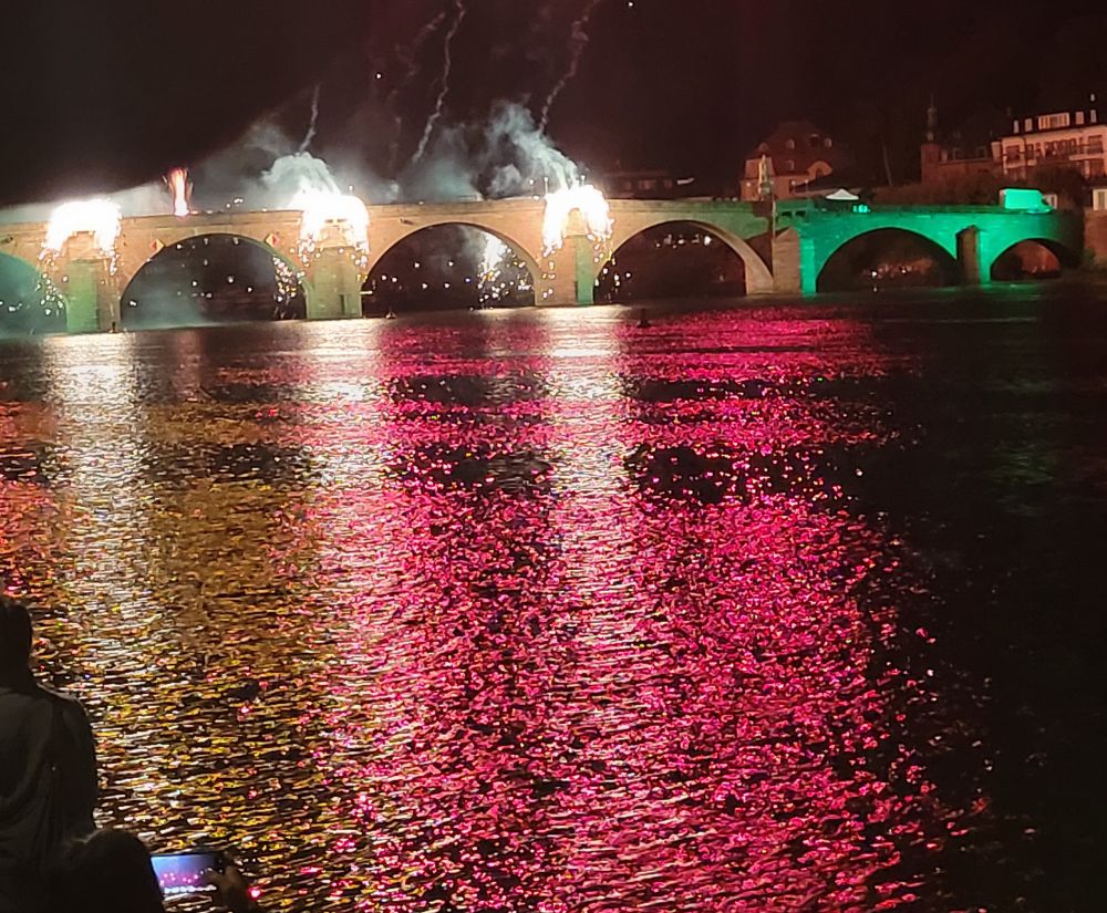 The old bridge of Heidelberg town lit by fireworks with the river Neckar appearing orange and crimson in the reflection.