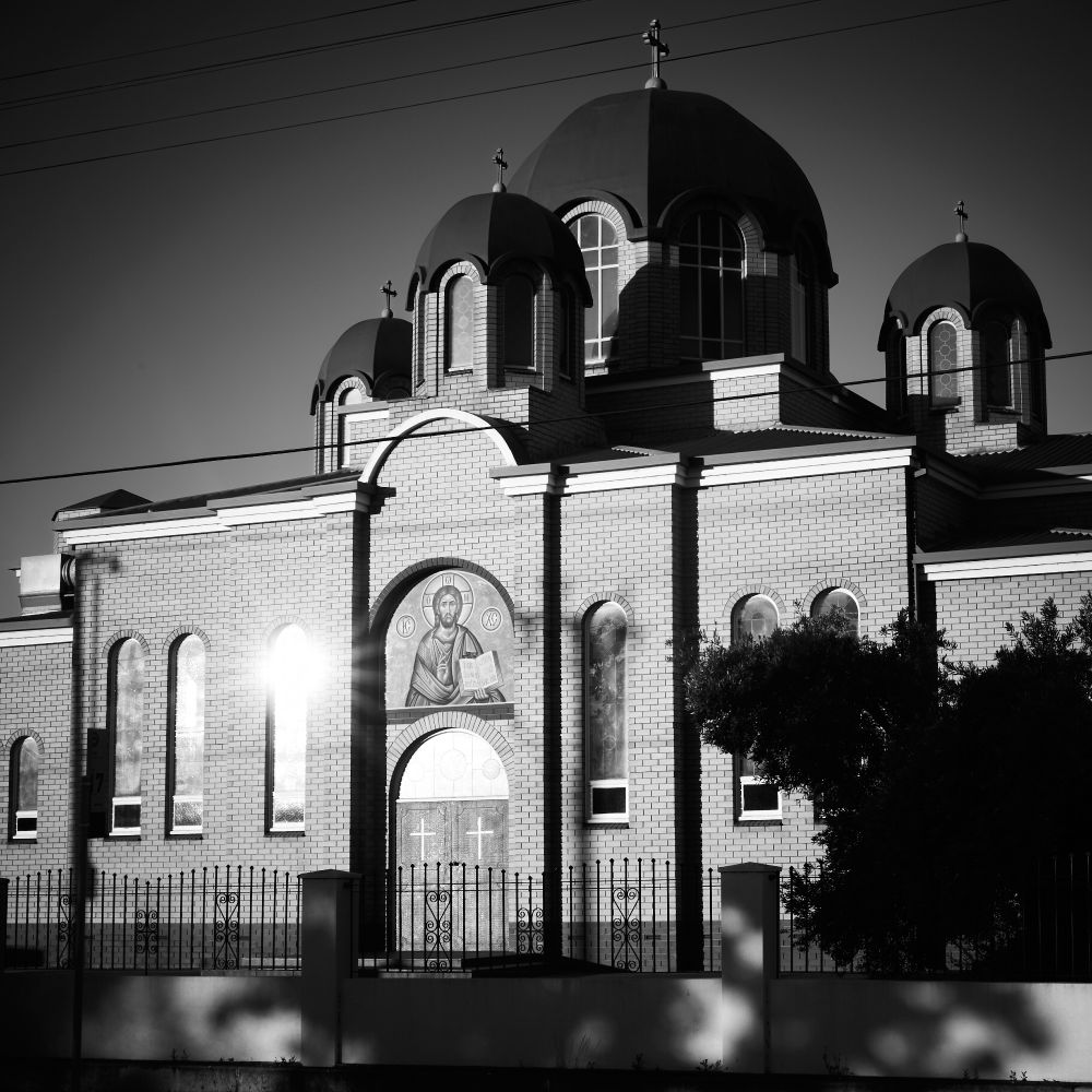 A christian church with domed roofs and an icon of Jesus above the door. It's lit from the side in late afternoon light, which is glinting off of the windows.