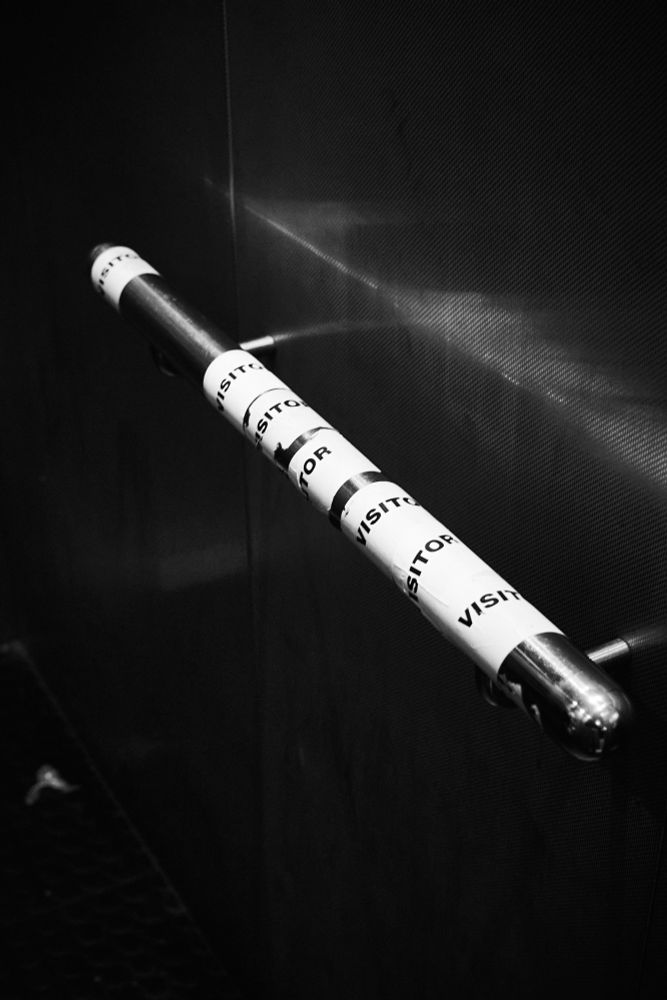 A black and white photograph of a handrail in an elevator, hospital visitor stickers discarded upon it.