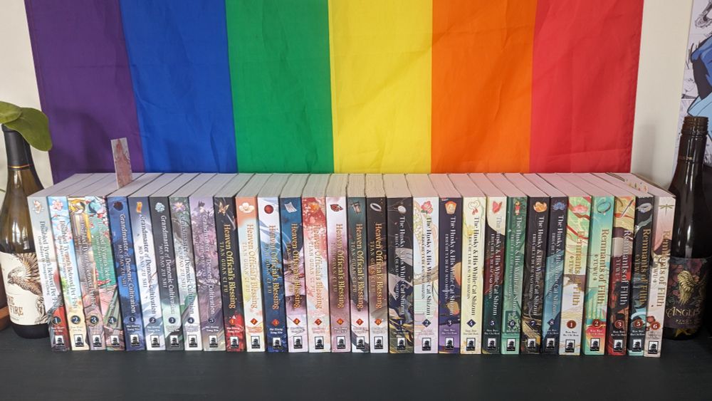 Shelf with my Danmei book collection including The Disabled Tyrant's Beloved Pet Fish, Grandmaster of Demonic Cultivation, Heaven Official's Blessing, The Husky and His White Cat Shizun, and Remnants of Filth (and a pride flag in the background)