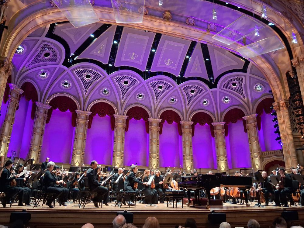 The Vancouver Symphony Orchestra warming up before the Friday evening concert. Strings in front row. All members wear black. A Steinway grand piana awaits guest pianist Zlata Chochieva.