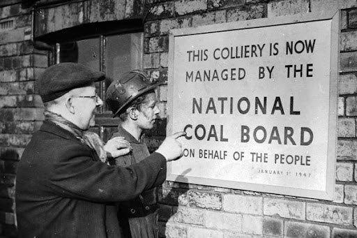 Black and white photo, two men examine a sign saying "This colliery is now managed by the national coal board on behalf of the people"