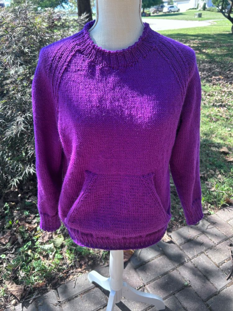 Bright purple, long sleeved pullover with a front pocket on a dress form with bricks and a mini Japanese maple for the backdrop.