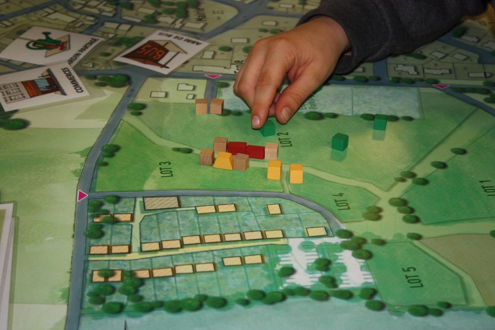 Un atelier d’urbanisme pour les petits. On voit la main d’un enfant qui saisit des cubes de bois à placer sur un plan de quartier d’une ville imaginaire.