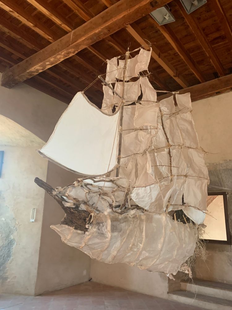 Un bateau en papier froissé et en fines tiges de bois semble s’envoler. Il est suspendu à un plafond en forme de coque renversée.