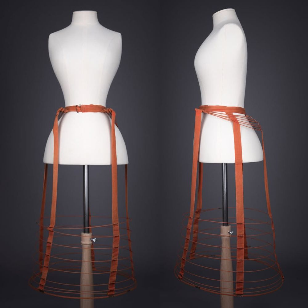 Date: c. 1870s

Origin: Great Britain

Fabric: Cotton tape, metal wires, metal buckle

Brand: Unknown

 

A cage crinolette of cotton tape, supported by textile encased metal wires and fastening at the front of the body with a metal buckle.

Crinolines offered support to the skirts of outer clothing whilst remaining lightweight and allowing a greater degree of freedom of movement. Previously, fashionable full skirted fashions were supported by a number of petticoats, which could be bulky and heavy. Cage crinolines were first popularised in the late 1850s, with the silhouette evolving to suit changing womenswear fashions. By the late 1860s, the full rounded crinoline shape had evolved into the crinolette, a transitional garment between the original cage crinoline and the bustles of the late 19th century. Crinolettes had a greater level of projection to the rear of the garment, with little projection to the sides and front of the garment.
