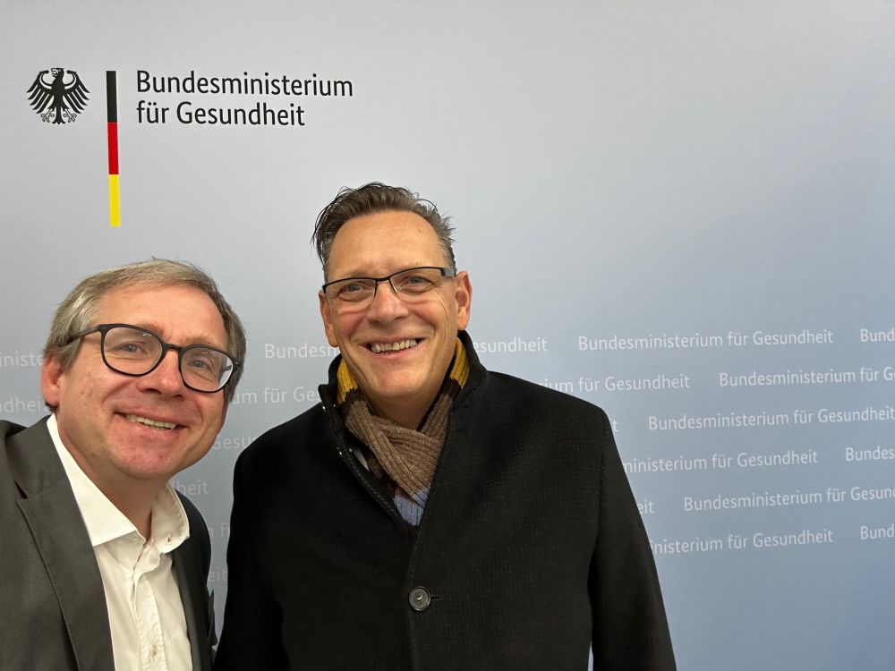 Carsten Müler-Tidow and Frank Buchholz smiling to the camera in front of the ministry's photo board.