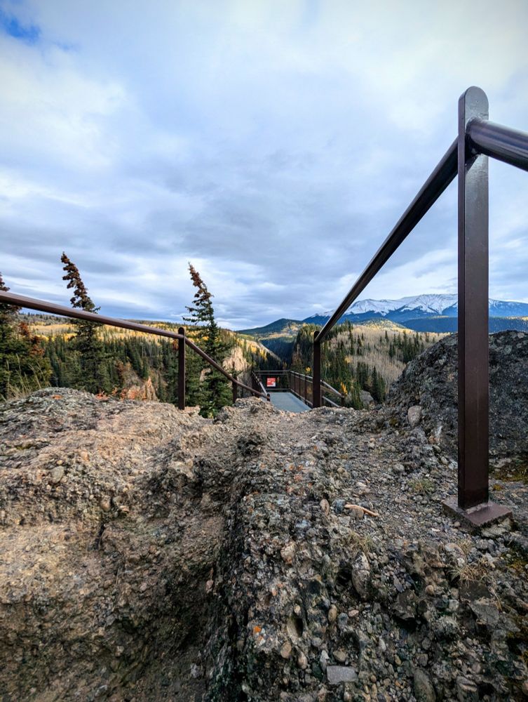 A large boulder with metal-gated sides leading to a lookout on the edge of a cliff with a view of mountains beyond; some covered in trees at various stages of Fall foliage, and beyond that some snow capped. A partly cloudy day. 
Sulphur Gates, Alberta, Canada. Oct 5, 2025.