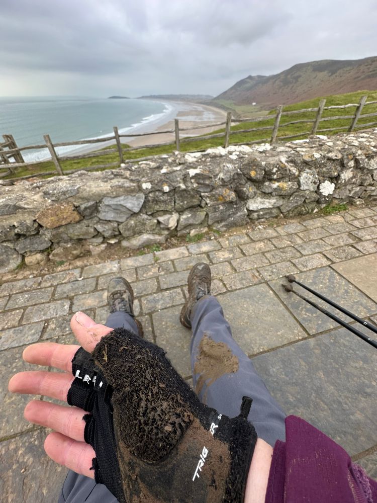 Muddy glove-covered hand with someone’s legs in muddy trousers and walking boots in the foreground. In the background is a curved bay showing sea, beach and hill.