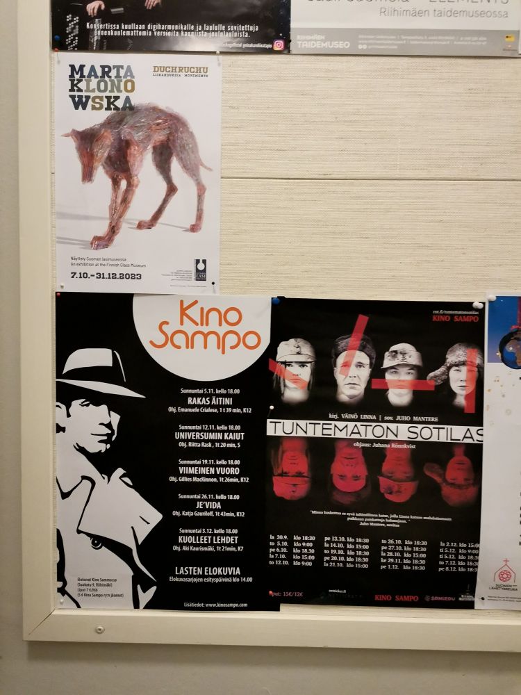 Seinällä ilmoitustaulu, jossa Suomen Lasimuseon ja Kino Sammon mainosjuliste. Kino Sampo ilmoittaa elokuvanäytöksistä ja Riihimäen Nuorisoteatterin "Tuntematon sotilas" -näytelmästä.