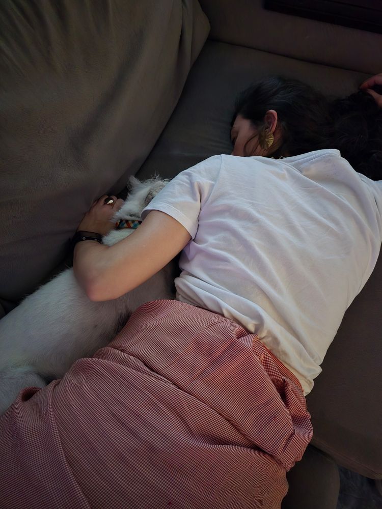 Mulher de camiseta branca e saia vermelha dormindo abraçada em cachorrinho branco