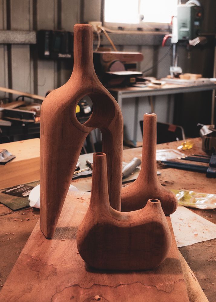 Three bud wooden bud vases on a wood workshop bench. The one at the front has two necks one short and one long, the middle one has long triangular legs with a hole through the body and a tall neck and the back one has a offset tall neck on a squat round body.