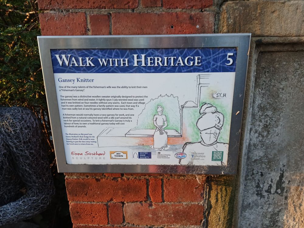 Photo of interpretation board for Gansey Knitter, sculpture 5 on the Walk with Heritage. The board includes design sketches and sponsor logos 