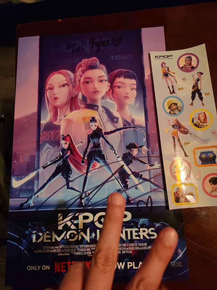 A signed promotional poster for K-Pop Demon Hunters that has "To: Tyler" written on top with EJAE's signature going across the bottom. A sticker sheet of a bunch of the characters is to the right and a hand is coming out of the bottom of the shot making a peace sign.