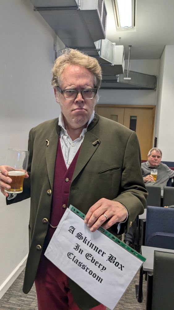 I am standing in the side of a lecture theatre. I wear glasses, my greying sideburns are fluffed and I have a stern expression. I'm wearing a dark green jacket, maroon waistcoat with a watch fob. In one hand a half finished beer and the other, a printed sign with gothic text reading "a Skinner box in every classroom". 
In background behind me an oblivious Prof Howell sits with a beer of his own