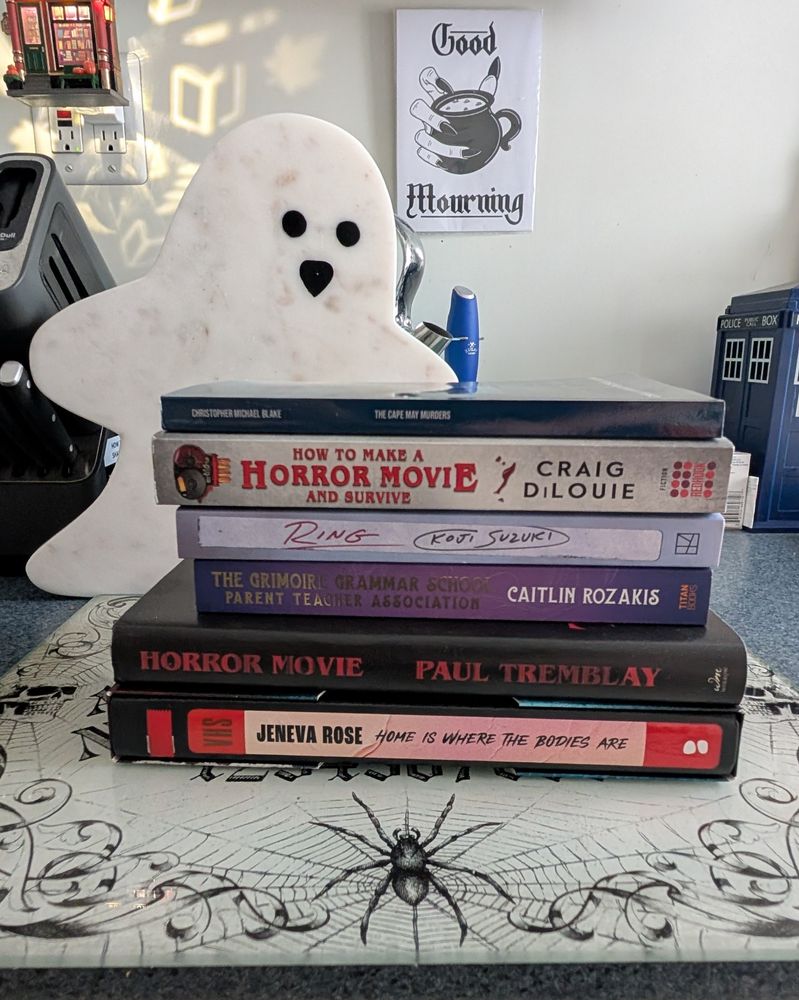 A stack of books on top of a stylized Ouija board. Behind it stands a granite ghost. On the wall is a drawing of a witch hand holding a small cauldron with the text "Good Mourning." There's a small Tardis off to the right. The books are (top to bottom) The Cape May Murders by Christopher Michael Blake, How To Make a Horror Movie and Survive by Craig DiLouie, Ring by Koji Suzuki, The Grimoire Grammar School Parent Teacher Association by Caitlin Rozakis, Horror Movie by Paul Tremblay, and Home Is Where the Bodies Are by Jeneva Rose. 