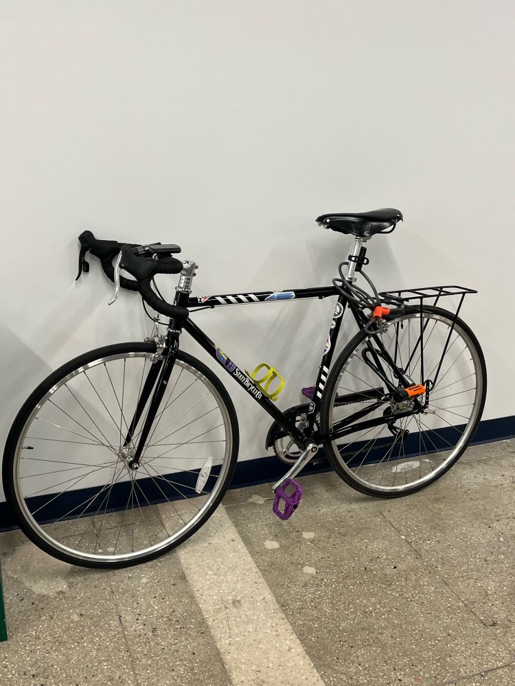 Black road bike with stickers and purple pedals, leaning against a wall.