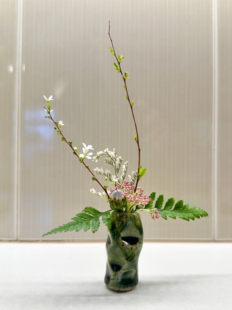 Miniature Sogetsu ikebana floral arrangement with flowers and blooming branches in a long green vase. 